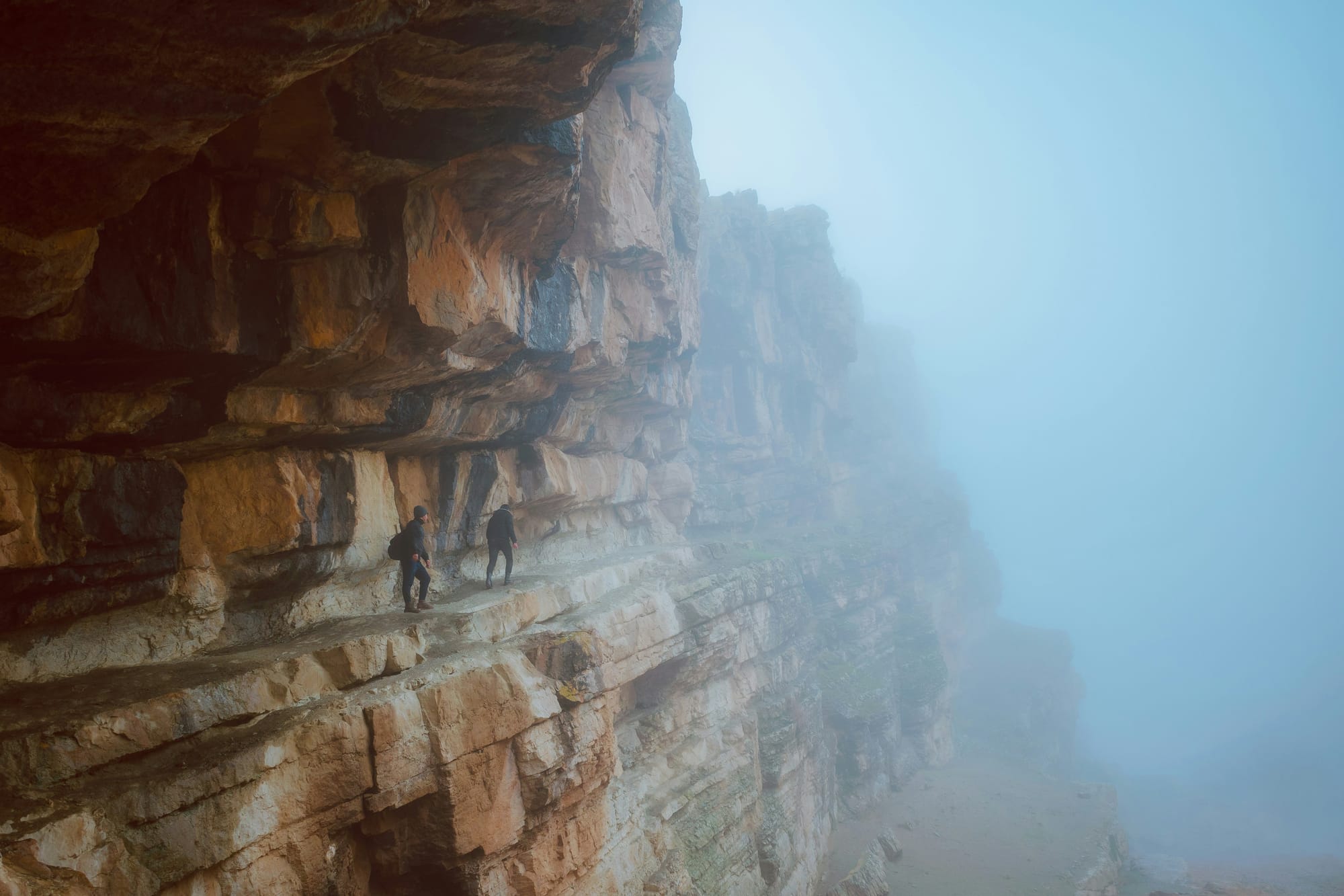 Two climbers traversing a narrow rocky ledge on a cliff face, with fog obscuring the drop below