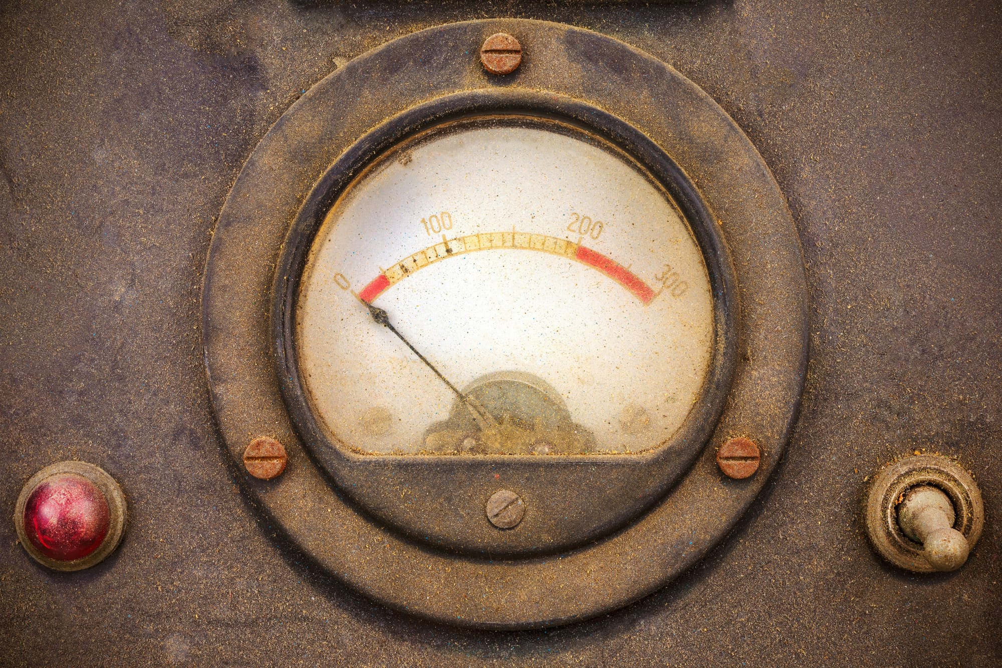 A weathered analog meter with a red zone marked on the dial, a small red indicator light to the left and a toggle switch to the right