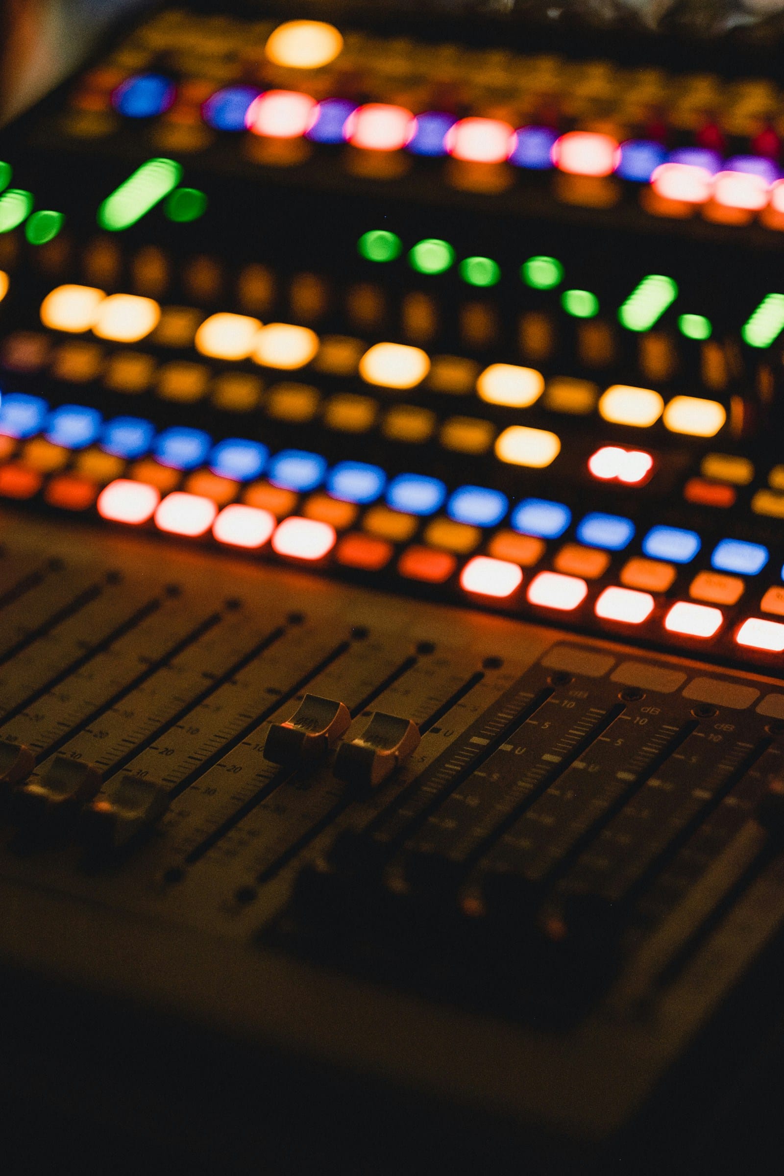 A mixing desk in the dark, rows of faders beneath glowing coloured buttons