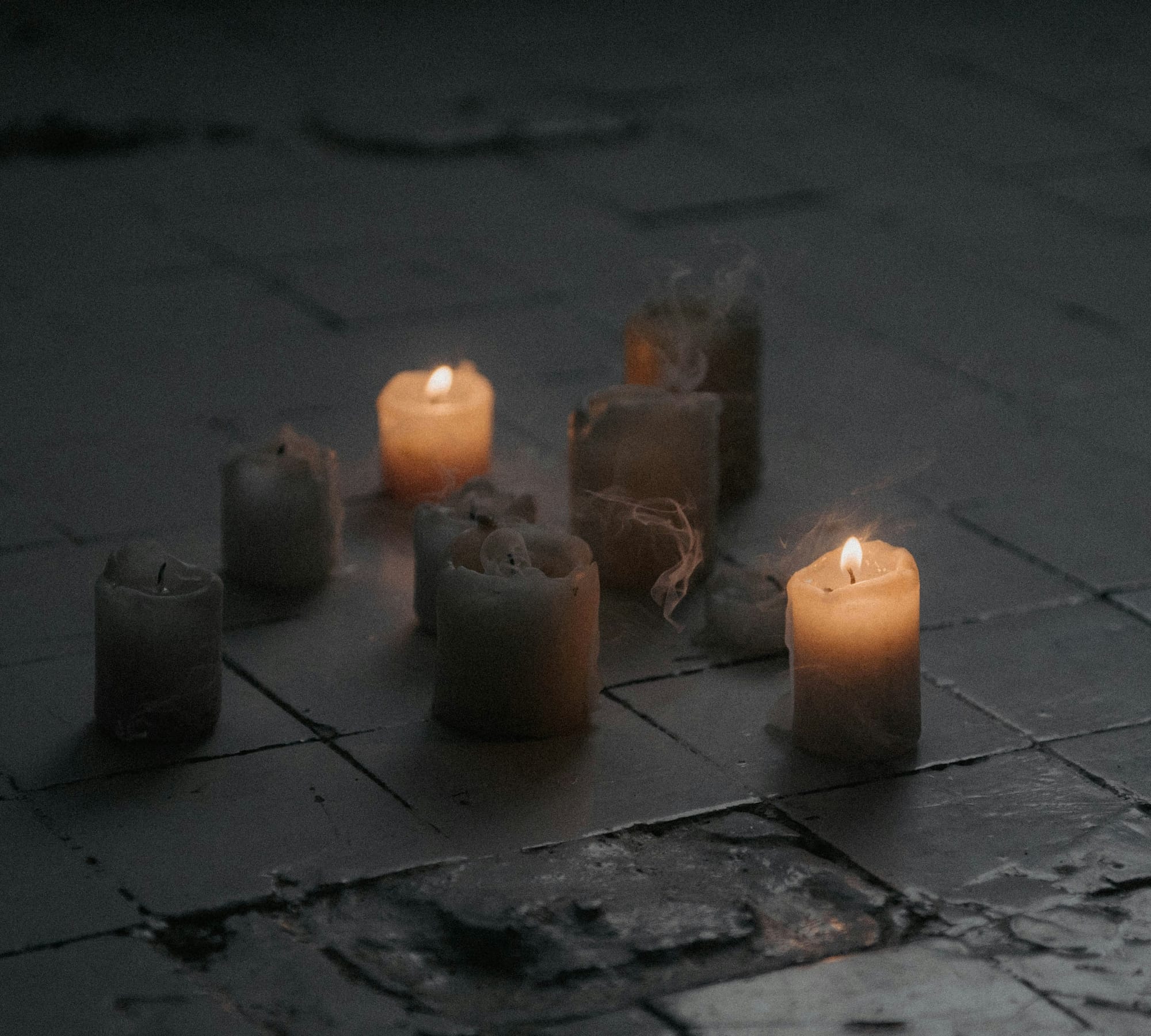 Candles on a dark stone floor, most of them burned out and trailing smoke, one or two still lit