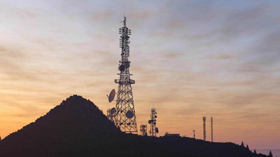 Silhouette of a telecommunications tower with antennas and dishes against a soft sunset sky, smaller towers on the horizon
